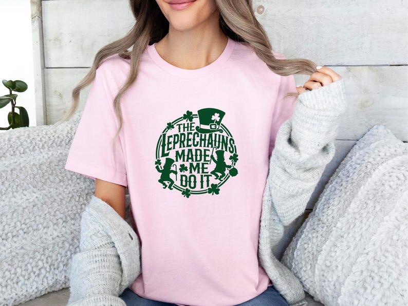 May include: A light pink t-shirt with a green graphic that says "THE LEPRECHAUNS MADE ME DO IT." The design includes a leprechaun, a hat, and shamrocks. The shirt is a crew neck style.