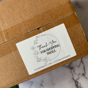 May include: A white sticker with the text "Thank You For Shopping Small" in a black script font, surrounded by a floral wreath. The sticker is affixed to a brown cardboard box.