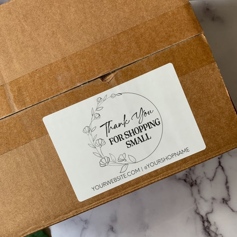 May include: A white sticker with the text "Thank You For Shopping Small" in a black script font, surrounded by a floral wreath. The sticker is affixed to a brown cardboard box.