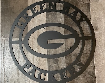 Puede incluir: Señal de metal negro con las palabras "Green Bay Packers" en un círculo alrededor de una gran "G" en el centro.