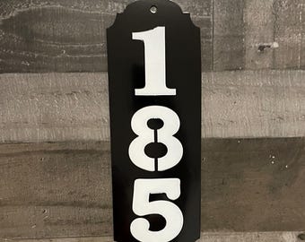 Pode incluir: Placa de número de casa de metal preto com números brancos 185. A placa tem uma borda decorativa.