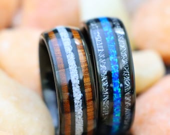 May include: Two black rings with unique designs. One ring features a wood grain and white stripe pattern, while the other has a blue and white opal-like inlay. The rings are set against a background of small, colorful stones.