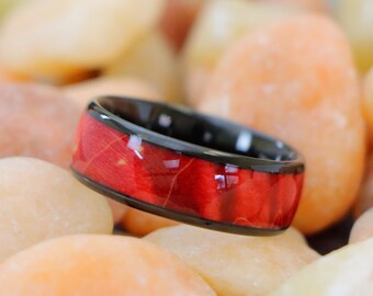 May include: A black and red ring with a wood inlay. The ring is sitting on a bed of small, smooth stones.