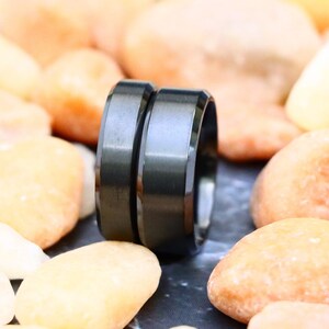 May include: Two black wedding bands with a brushed finish. The bands are stacked on top of each other and are sitting on a bed of small, light brown stones.