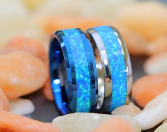 May include: Two wedding bands with blue and silver inlay. The bands are made of tungsten carbide and feature a blue opal inlay. The bands are sitting on a bed of orange and white stones.