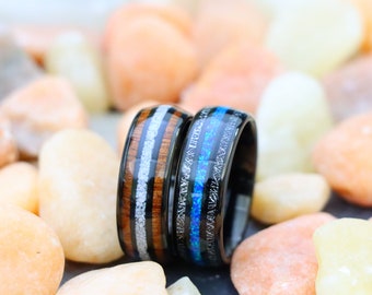 May include: Two black tungsten carbide rings with different inlays. One ring has a wood and white inlay, the other has a blue and white inlay.