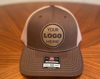 Könnte beinhalten: Braune und beigefarbene Trucker-Kappe mit einem braunen Patch, auf dem "YOUR LOGO HERE" steht. Die Kappe ist verstellbar und hat ein Richardson 112-Etikett.