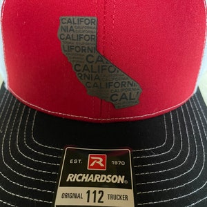 Könnte beinhalten: Rote und weiße Trucker-Mütze mit schwarzem Schirm. Die Mütze zeigt eine Umrisslinie des Bundesstaates Kalifornien mit dem Wort "California" innerhalb der Form. Die Mütze hat ein Richardson-Etikett.