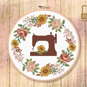 Può includere: Un cerchio da ricamo bianco con un design a corona di fiori con rose rosa, girasoli gialli e foglie verdi. Una silhouette marrone di una macchina da cucire è centrata nella corona.