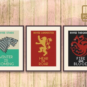May include: Three framed cross-stitch patterns featuring the sigils of House Stark, House Lannister, and House Targaryen from the TV show Game of Thrones. The Stark sigil is a grey direwolf, the Lannister sigil is a golden lion, and the Targaryen sigil is a three-headed red dragon. The text below each sigil reads "Winter is Coming", "Hear Me Roar", and "Fire and Blood" respectively.