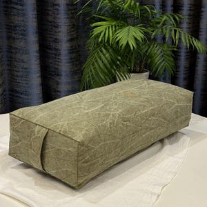 Puede incluir: Un cojín de yoga rectangular verde oliva con una tela texturizada y arrugada. El cojín tiene un asa en un lado y se coloca sobre una superficie blanca. Una planta en maceta con hojas verdes está en el fondo.