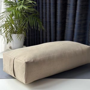 Puede incluir: Un cojín de yoga rectangular en color beige neutro. El cojín está hecho de una tela suave y tiene un diseño simple y minimalista. Se coloca sobre una esterilla de yoga gris, con una planta en maceta y cortinas azules en el fondo.