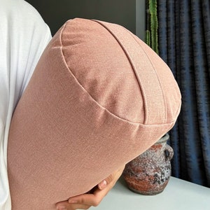 Könnte beinhalten: Ein rosa Yoga-Kissen aus weichem, strukturiertem Stoff. Das Kissen hat eine abgerundete Form und ist perfekt, um den Körper während Yoga-Posen zu stützen.