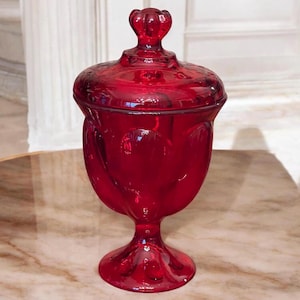 May include: A red glass candy dish with a lid and a pedestal base. The dish has a textured design and a rounded shape.