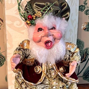 May include: A whimsical Christmas elf figurine with a rosy face, white beard, and open mouth. The elf wears a green hat adorned with berries and greenery, and a gold and burgundy outfit with bells and pearl accents. The figurine is a festive holiday decoration.