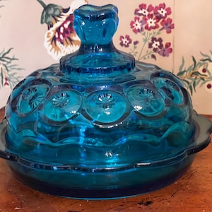 May include: A vintage, teal-colored glass butter dish with a lid. The lid features a decorative handle and a pattern of starbursts. The dish has a scalloped edge and sits on a round base. The background is a floral wallpaper.