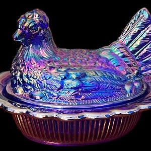May include: Iridescent blue hen-shaped covered dish. The dish features a detailed hen with a textured surface and a ruffled edge. The dish is made of glass and has a vibrant, multi-colored sheen.
