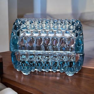 May include: A blue glass trinket box with a textured, bubble-like design. The box has a hinged lid and is made of clear glass with a light blue tint.