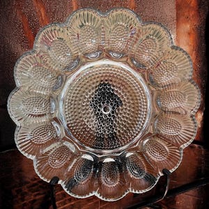 May include: Clear glass serving dish with a scalloped edge and a central circular section with a textured pattern. The outer edge has oval-shaped indentations. The dish is designed to hold eggs or other small items.