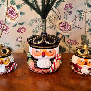 May include: Three Christmas ornaments shaped like nutcrackers. Each ornament has a gold crown, a black hat, and a red and white uniform. The nutcrackers are holding candy canes. The ornaments are on a wooden surface.