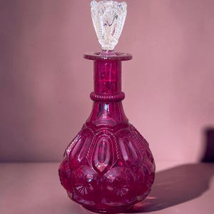 May include: A deep magenta glass decanter featuring a bulbous body with a repeating embossed oval and starburst pattern. The neck has a ribbed collar, and the top is finished with a clear, faceted glass stopper that has a pointed, petal-like design.