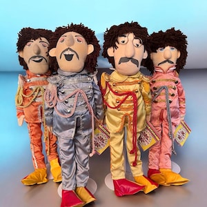 May include: Four plush dolls dressed in colorful military-style jackets and pants, each with a unique color scheme: orange, silver, yellow, and pink. The dolls have brown curly hair, mustaches, and are holding small cards.