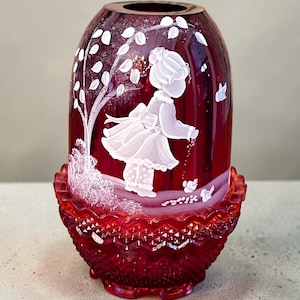 May include: A ruby red glass vase with a white painted design of a girl, tree, and butterflies. The vase has a flared, textured base and a rounded top. The design is hand-painted, adding a unique touch to the decorative piece.