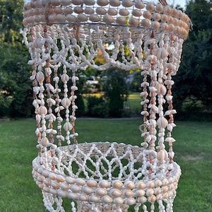 May include: A white seashell chandelier with a wire frame and a woven design. The chandelier is hanging from a rope and is decorated with seashells.