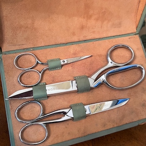 May include: A set of three silver-toned metal scissors with polished blades and finger loops, secured by sage green leather straps inside a tan leather-lined case. The blades are engraved with the brand name Singer, and the case is marked Germany.
