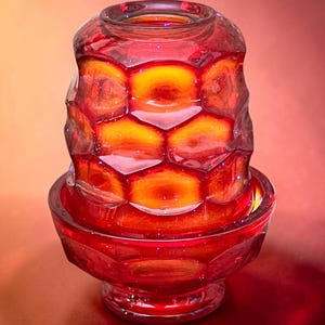 May include: A deep red glass candle holder featuring a textured honeycomb pattern on the upper section and a smooth, circular base. The translucent material catches the light, creating a gradient of vibrant crimson and orange tones throughout.