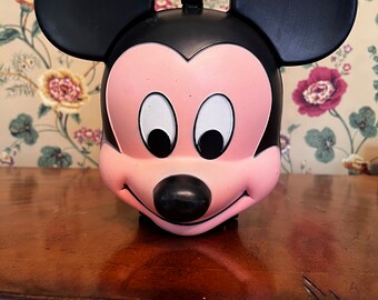 May include: A pink and black plastic Mickey Mouse lunchbox with a handle. The lunchbox has a smiling face with black eyes and a black nose.