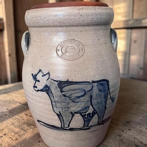 May include: A beige stoneware crock with a dark blue hand-painted cow illustration on the front. The vessel features two side handles, a brown wooden lid, and a circular stamp that reads Country Home Collection with a house icon in the center.