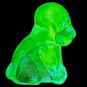 May include: A bright green glass figurine of a seated dog, set against a black background. The dog has a rounded head, floppy ears, and a small tail. The glass has a slight texture and a heart-shaped marking.