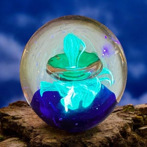 May include: A glass paperweight with a blue, green, and white swirling design inside. The paperweight is round and clear, and the design is visible from all sides.