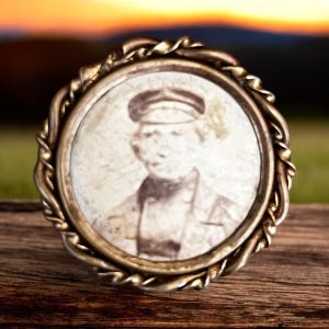 May include: A vintage brass locket with a woven rope design around the edge. The locket contains a faded black and white photograph of a person wearing a cap.