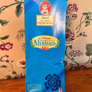 May include: A rectangular blue toy box featuring an illustration of Ariel from Disney's The Little Mermaid. The box displays the text Ariel Sea Pearl Princess, Disney's The Little Mermaid, and a warning label about small parts at the bottom corner.
