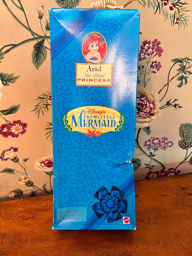 May include: A rectangular blue toy box featuring an illustration of Ariel from Disney's The Little Mermaid. The box displays the text Ariel Sea Pearl Princess, Disney's The Little Mermaid, and a warning label about small parts at the bottom corner.