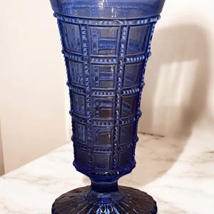 May include: A cobalt blue glass vase with a textured, geometric pattern. The vase has a wide, fluted top and a pedestal base. The design features a series of squares and rectangles, creating a grid-like appearance. The vase is approximately 10 inches tall.