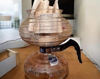 Puede incluir: Una cafetera de vidrio vintage con un asa negra y una base plateada. La cafetera tiene una base redonda y una parte superior más ancha, llena de papeles enrollados. La etiqueta dice "VisiVac".