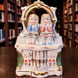 May include: A vintage porcelain figurine featuring two young girls seated on a decorative bench. The figurine has a white base with gold trim and accents of green and red. The girls are dressed in blue and pink outfits.