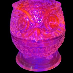 May include: A decorative, glass owl-shaped container with a lid. The container is a vibrant orange and purple color, with the owl's features, including large eyes and a beak, clearly visible. The container sits on a clear, circular base.