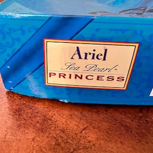 May include: A blue rectangular cardboard box featuring a prominent white label with a gold border. The text on the label reads Ariel Sea Pearl PRINCESS in black and red lettering. The box surface has a decorative blue pattern and branding.
