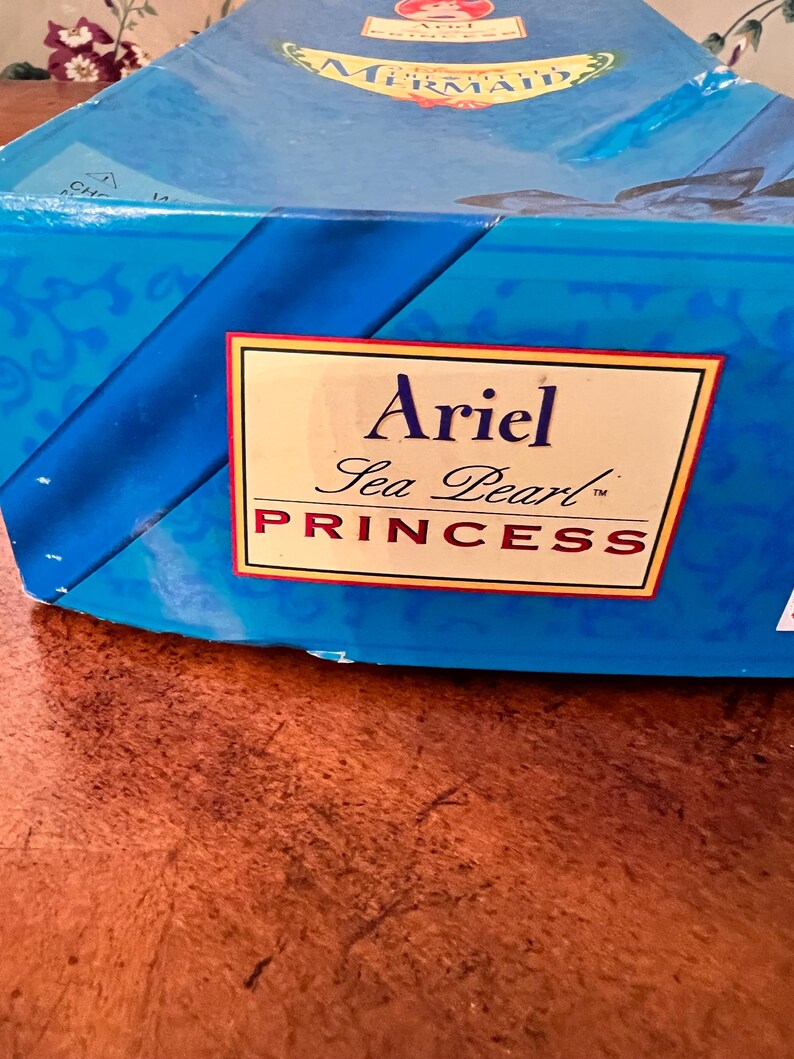 May include: A blue rectangular cardboard box featuring a prominent white label with a gold border. The text on the label reads Ariel Sea Pearl PRINCESS in black and red lettering. The box surface has a decorative blue pattern and branding.