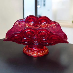 May include: A red glass bowl with a scalloped edge and a pedestal base. The bowl has a textured surface with small raised circles.
