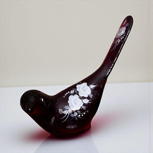 May include: A burgundy glass bird figurine with white floral accents. The bird has a long, curved neck and a rounded body. The white flowers are painted on the body and neck of the bird. A decorative item.