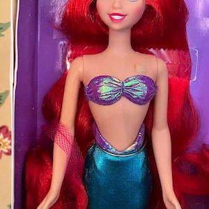 May include: A mermaid doll with long, vibrant red hair, blue eyes, and a light skin tone. The doll wears a purple iridescent seashell-style top and a metallic teal mermaid tail. The figure is positioned inside a purple box with visible packaging.