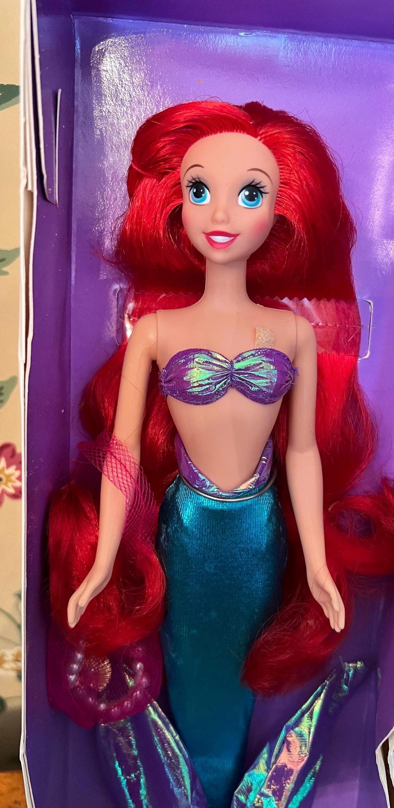 May include: A mermaid doll with long, vibrant red hair, blue eyes, and a light skin tone. The doll wears a purple iridescent seashell-style top and a metallic teal mermaid tail. The figure is positioned inside a purple box with visible packaging.