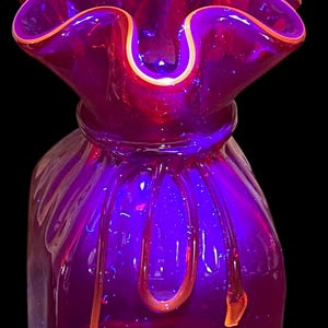 May include: A vibrant, purple glass vase with a ruffled rim and a unique, textured design. The rim is outlined with a thin orange border. The vase has a bulbous shape with vertical lines and orange accents.
