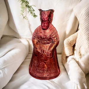May include: A vintage glass pitcher shaped like a woman wearing a long dress. The pitcher is made of pink glass and has a textured surface.