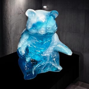 May include: A translucent blue bear sculpture with a sitting pose. The bear's form is detailed, with visible textures and a gradient of blue hues. The sculpture is displayed on a black surface against a dark gray background.